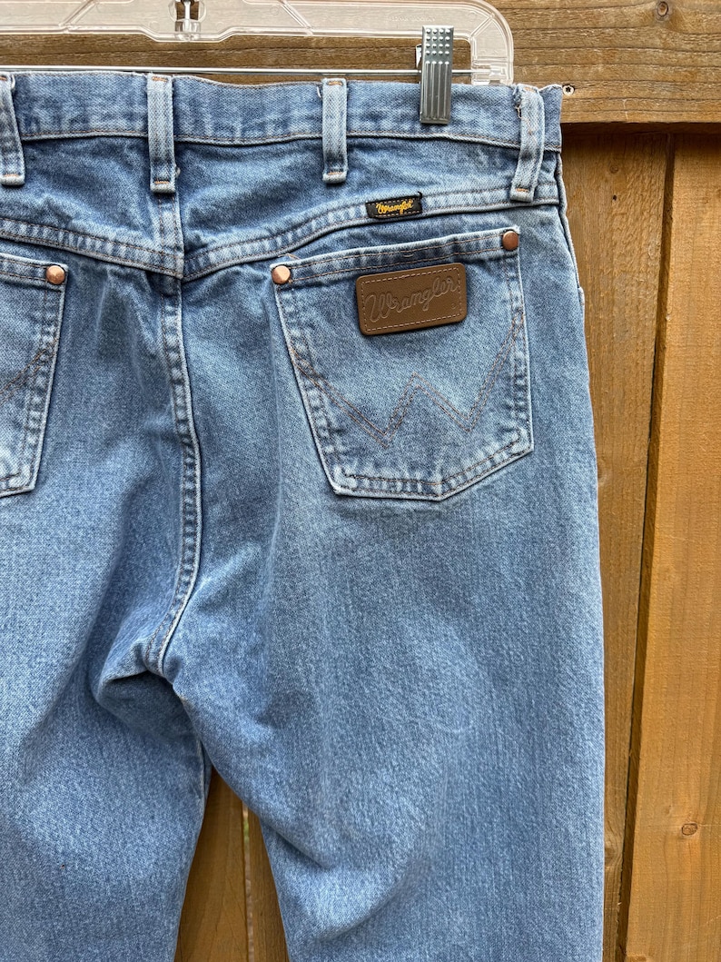 May include: Light blue denim jeans with copper rivets and a brown leather Wrangler patch on the back pocket. The jeans are hanging on a metal hanger. The Wrangler logo is visible on the patch and above the back pocket.