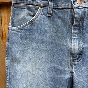 May include: Close-up of light blue denim jeans. The jeans have a classic five-pocket design, with a button and zipper closure. The fabric shows subtle fading and wear, suggesting a vintage or well-loved item.