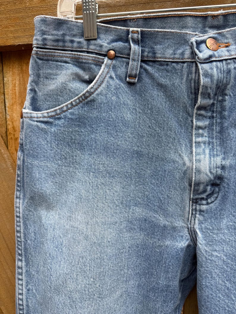 May include: Close-up of light blue denim jeans. The jeans have a classic five-pocket design, with a button and zipper closure. The fabric shows subtle fading and wear, suggesting a vintage or well-loved item.