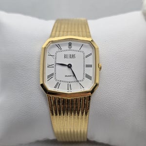 May include: A gold-toned Bill Blass quartz watch with a rectangular face and a gold mesh band. The watch face is white with black Roman numerals and hands. The watch is displayed on a white pillow.