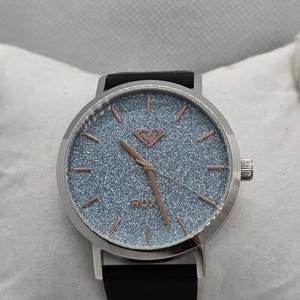 May include: A silver-toned wristwatch with a round face and a black leather band. The watch face has a light blue glittery background with rose gold-colored hands and markers. The brand name "ROXY" is visible on the watch face.