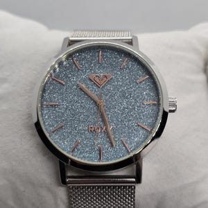 May include: A silver-toned Roxy watch with a shimmering blue-gray face and rose gold hands and markers. The watch features a mesh band and a heart-shaped logo. The watch is a stylish accessory.