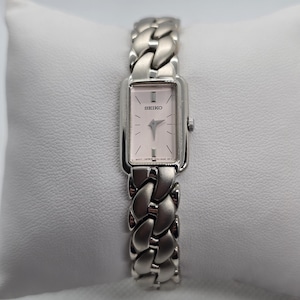 May include: A silver-toned Seiko wristwatch with a rectangular face and a braided metal band. The watch face is a light pink color with silver hands and markers. The watch is displayed on a white pillow.
