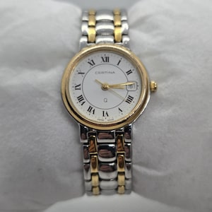 May include: A Certina wristwatch with a gold-tone bezel and accents, featuring a white face with Roman numeral hour markers. The watch has a date display at the 3 o'clock position and a two-tone stainless steel and gold-tone band.