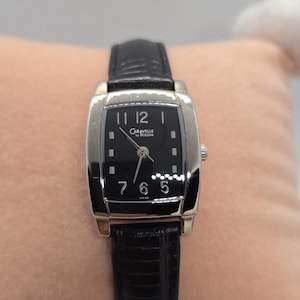 May include: A Caravelle by Bulova wristwatch with a rectangular black face and silver-tone case. The watch has a black leather band and displays the time with silver hands and numerals. The watch is a classic accessory.