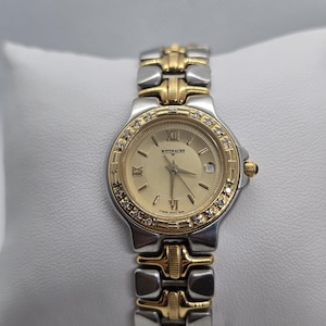 May include: A gold and silver Wittnauer wristwatch with a gold dial, Roman numeral hour markers, and a date display at the 3 o'clock position. The bezel is adorned with small, clear stones. The watch has a two-tone metal band.