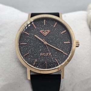 May include: A Roxy wristwatch with a black glittery face and rose gold accents. The watch has a gold-colored bezel and a black band. The Roxy logo is visible on the watch face. The watch has a simple, elegant design.