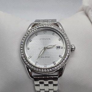May include: A Citizen Eco-Drive watch with a silver band and a white face. The watch face is surrounded by a ring of small, clear stones. The watch has a date display at the 3 o'clock position.