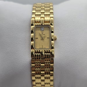 Puede incluir: Un reloj de pulsera Bulova dorado con una esfera rectangular y un diamante. El reloj tiene una esfera texturizada y una pulsera de eslabones. El reloj se muestra sobre una superficie blanca.