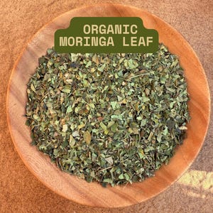May include: A wooden bowl filled with dried green moringa leaves. A green label with white text that reads "ORGANIC MORINGA LEAF" is placed above the leaves. The leaves are a mix of green and brown.