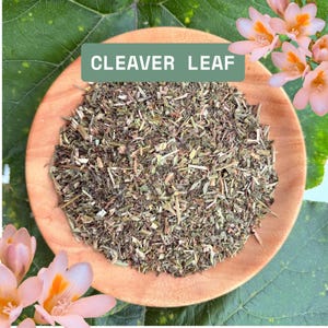 May include: A round wooden bowl filled with dried, chopped green herbs labelled CLEAVER LEAF. The bowl is surrounded by large green leaves and small, light pink flowers with orange centres, creating a natural, botanical display of the herb.