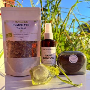 May include: A selection of natural wellness products, including a tea blend, body oil, and a mesh tea infuser. The tea blend is in a white pouch with text that reads "LYMPHATIC Tea Blend". The body oil is in a brown bottle with a spray top.