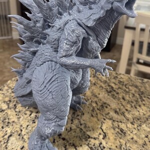 May include: A gray 3D printed model of Godzilla, a fictional monster, with a detailed textured surface and sharp spikes on its back. The model is posed in a standing position with its mouth open and its claws extended.