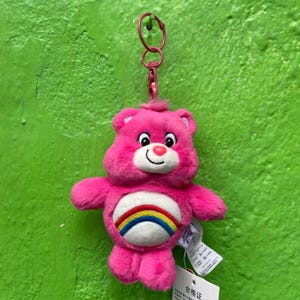 May include: A pink Care Bear plush keychain with a rainbow belly and a smiling face. The plush toy has a pink metal clasp and a small tag attached. The background is a solid green colour.