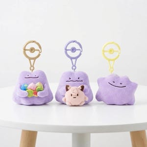 May include: Three plush keychains featuring purple Ditto characters with different expressions. One holds colorful items, another has a small pink character, and the third is star-shaped. Each keychain has a different colored clip.