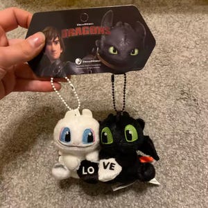 May include: Two plush dragon keychains, one white and one black, with the word "LOVE" on a heart. The white dragon has blue eyes, and the black dragon has green eyes. A DreamWorks Dragons tag is attached.