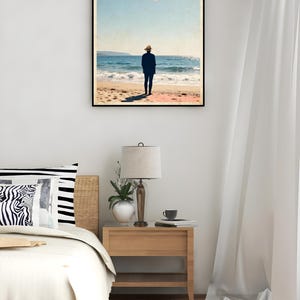 Puede incluir: Un póster enmarcado con el texto "Mediterranean Escape" en rosa. La imagen muestra a una persona en una playa, mirando al mar. El póster está en un dormitorio, junto a una cama y una mesita de noche con una lámpara.
