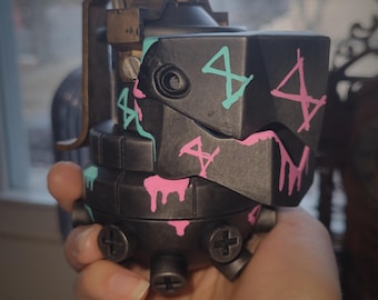 May include: A black grenade-shaped prop featuring a metallic finish with protruding screw-like details at the base. The surface is decorated with hand-painted teal and pink geometric symbols and drip patterns, with a dark metal lever on the side.