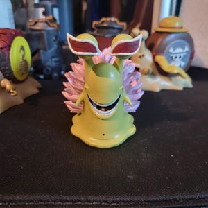 Puede incluir: Una figura amarilla de una criatura parecida a un caracol con detalles rojos y blancos en la cabeza y una voluta rosa. La figura tiene una cara sonriente y forma parte de una colección de figuras similares.