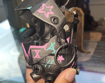 May include: A dark gray, mechanical device with pink and teal graffiti-style markings, including stars and cross shapes. The device has a ring and a lever, suggesting a functional or decorative item. The design is detailed and industrial.