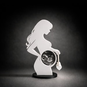 Könnte beinhalten: Eine weiße Silhouette einer schwangeren Person, die eine Babyrassel hält. Die Silhouette hat einen Ausschnitt, der ein Ultraschallbild zeigt. Die Figur ist auf einem schwarzen kreisförmigen Sockel montiert.