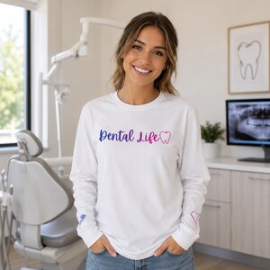 May include: A white long-sleeve shirt featuring the text Dental Life in a blue to pink gradient font, accompanied by a small tooth icon. The right sleeve displays a small blue toothbrush graphic, and the left sleeve features a small purple heart.