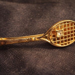 May include: A gold-colored tennis racket brooch with a diamond-like center stone. The brooch has a detailed, textured handle and a grid-like pattern on the racket head. The brooch is set against a dark, textured background.