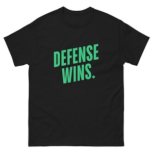 May include: White t-shirt with the text "DEFENSE WINS." in bold, green letters. The shirt has a crew neck and short sleeves. The text is angled slightly from the upper left to the lower right.