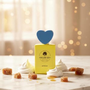 May include: A yellow perfume bottle with a blue heart-shaped cap, labeled "Willow Sky Parfum - Aspyn Haze." The bottle is surrounded by white meringue swirls and small, brown sugar cubes on a marble surface.