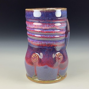 Puede incluir: Una taza de cerámica hecha a mano con un diseño único. La taza presenta una vibrante combinación de colores púrpura, rojo y azul. Tiene un cuerpo texturizado y acanalado y un asa. La base tiene un borde verde claro. Dos elementos decorativos están en la parte inferior de la taza.