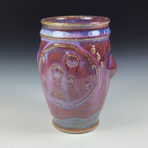 May include: A purple and brown ceramic mug with a textured surface and a circular design. The mug has a rounded bottom and a slightly flared top.