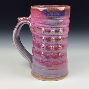 Puede incluir: Una taza de cerámica hecha a mano con un asa grande. La taza presenta una vibrante combinación de colores rosa, morado y dorado. El cuerpo tiene un diseño texturizado y acanalado con pequeños detalles dorados decorativos. La taza mide aproximadamente 15 cm de alto.