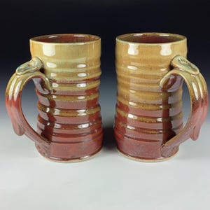 Puede incluir: Dos tazas de cerámica hechas a mano con un diseño acanalado. Las tazas tienen un degradado de colores, que va desde un beige claro en la parte superior hasta un rojo intenso en la base. Las asas tienen una forma y un color únicos, lo que aumenta el atractivo artístico.