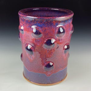 Puede incluir: Un vaso de cerámica hecho a mano con una superficie texturizada y un acabado brillante. El vaso es principalmente rojo con acentos morados y azules, y presenta una serie de decoraciones elevadas en forma de burbujas. La base es de un tono morado más oscuro.