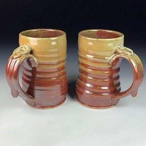 Puede incluir: Dos tazas de cerámica hechas a mano con un diseño rústico. Las tazas tienen forma cilíndrica con una textura acanalada y un asa grande y adornada. La combinación de colores incluye tonos marrones, beiges y rojos.