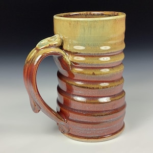 Puede incluir: Una taza de cerámica hecha a mano con un asa grande. La taza presenta un diseño acanalado en tonos marrones, rojos y amarillos. El asa tiene un elemento decorativo cerca de la parte superior. La taza es adecuada para bebidas frías o calientes.