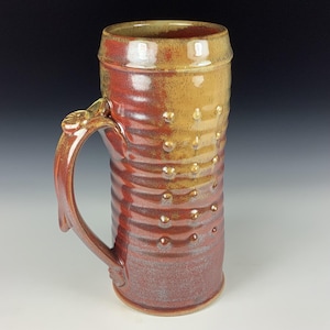 Puede incluir: Una jarra de cerveza alta de cerámica hecha a mano, con un asa. La jarra es principalmente roja y marrón con una superficie texturizada y pequeños puntos en relieve. La parte superior y el asa tienen un tono marrón dorado más claro.