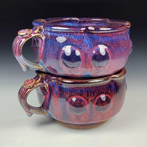 May include: Two stacked ceramic mugs with a whimsical design. The mugs are glazed in shades of purple, blue, and red, with a glossy finish. Each mug features a face-like design with large, round eye shapes. The handles have a decorative, floral-like detail.