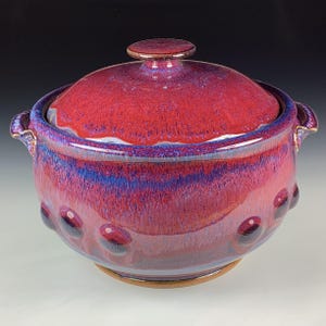 Puede incluir: Una fuente de cerámica hecha a mano con tapa. La fuente está esmaltada en tonos de rojo, morado y azul. Tiene forma redonda con pequeños elementos decorativos redondeados alrededor de la base. La tapa tiene un pequeño asa en la parte superior. La fuente mide unos 20 cm de diámetro.