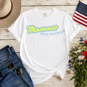 Puede incluir: Camiseta blanca con el texto "Mamaw" en fuente verde claro con contorno azul, y el texto "Jen's mockups" en azul. La camiseta está combinada con vaqueros azules, gafas de sol, un sombrero, flores y una bandera americana.