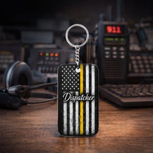 Könnte beinhalten: Ein rechteckiger Schlüsselanhänger mit einem Design der amerikanischen Flagge, einem gelben Streifen und dem Wort "Dispatcher". Er ist an einer silbernen Kette und einem Ring befestigt. Der Hintergrund ist verschwommen und zeigt Kommunikationsgeräte.