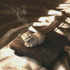 May include: A small, light-colored cat-shaped incense holder with a cigarette in its mouth, resting on a dark wooden platform. Wisps of smoke rise from the incense, creating a serene atmosphere. The platform sits on a textured, light-colored surface.