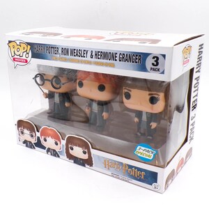 May include: A Funko Pop Movies 3-pack featuring vinyl figures of Harry Potter, Ron Weasley, and Hermione Granger. The white box displays the characters' names, the Harry Potter logo, and illustrations of the figures. The figures are visible through a clear plastic window, each wearing a dark school robe.
