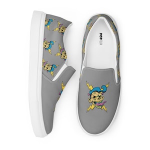 May include: A pair of gray slip-on canvas shoes with a white sole. The shoes feature a repeating pattern of a cartoon character with blue hair, a yellow face, and a pink tongue. The text "POPFELIX" is printed on the back of the right shoe.