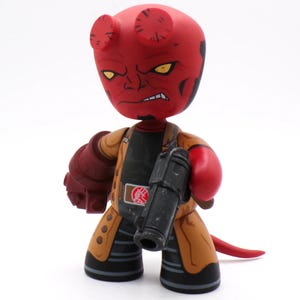 May include: A red Hellboy figurine with yellow eyes, horns, and a stern expression. The figure wears a tan coat, black shirt, and holds a black gun. The figure has a red tail and a large red right hand.