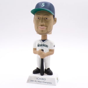 Puede incluir: Una figura de bobblehead de un jugador de béisbol que lleva una gorra de béisbol azul marino y verde y una camiseta blanca con "Mariners" impresa en la parte delantera. La figura sostiene un bate de béisbol negro y se encuentra sobre una base que dice "Ichiro 2001 AL West Champions."