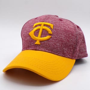 Puede incluir: Una gorra de béisbol con una corona de color granate jaspeado y una visera amarilla sólida. La parte frontal de la gorra muestra un prominente logotipo amarillo bordado que consiste en las letras T y C entrelazadas en un diseño audaz.