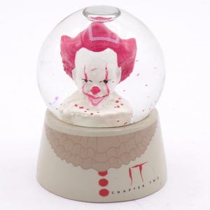 Puede incluir: Una bola de nieve de cristal transparente que contiene una figura de payaso con cara blanca, pelo rojo y marcas faciales rojas. La base es de color beige con un diseño de cuello con volantes y el texto rojo IT CHAPTER TWO en el frente.