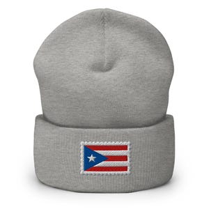 May include: A gray beanie featuring an embroidered Puerto Rican flag patch. The flag patch is rectangular with a blue triangle, a white star, and red and white stripes. The beanie is made of a soft, knitted material.