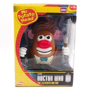 Puede incluir: Juguete de Mr. Potato Head en una caja, diseñado como el Undécimo Doctor de Doctor Who. El juguete tiene un cuerpo de patata marrón, una nariz roja y una pajarita azul. La caja es amarilla y roja con el logo de la BBC.